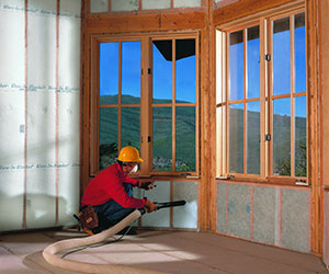 Worker installing blown-in insulation in a wall beneath windows.