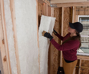 Worker installing white fiberglass batt insulation in a wall.
