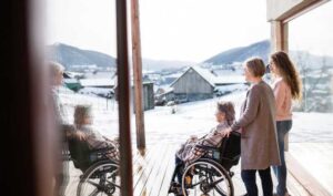 3 women on a porch in winter, with one seated in a wheelchair and covered with a blanket.
