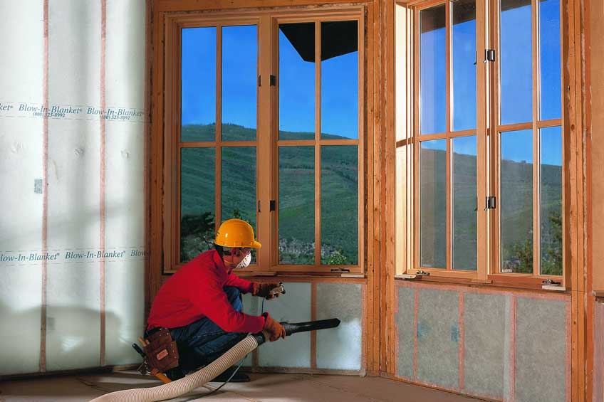 Worker installing blown-in insulation in a wall beneath windows.