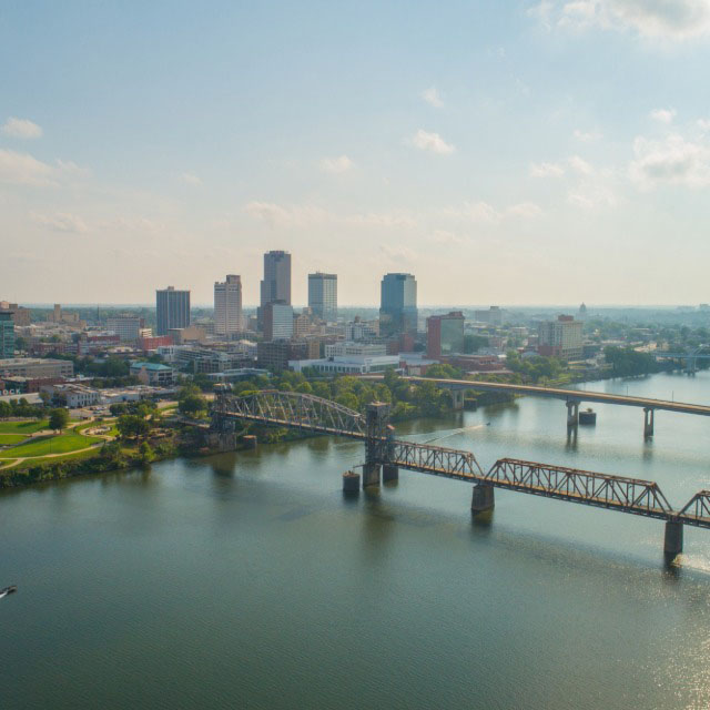An aerial view of the Little Rock, Arkansas skyline under a partly cloudy sky, featuring several high-rise buildings and multiple bridges crossing the wide Arkansas River in the foreground.